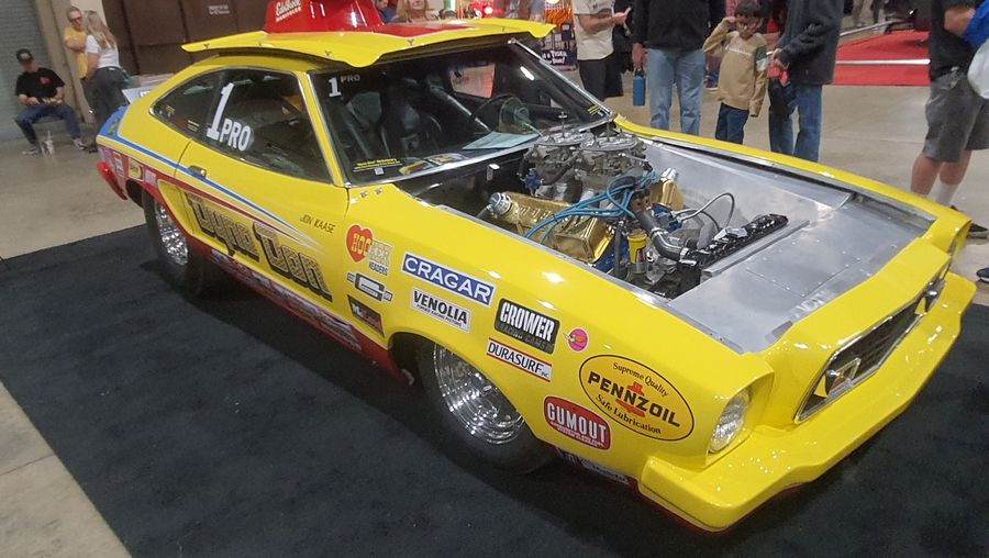 Żółty Ford Mustang II Pro Stock z lat 70. – drag car z naklejkami sponsorów i silnikiem bez maski w hali